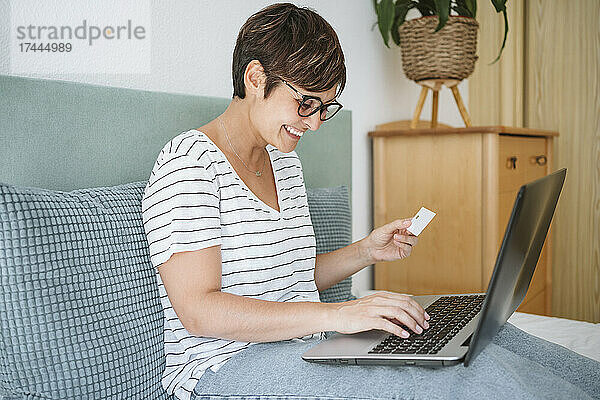 Glückliche Frau  die kontaktlos per Kreditkarte bezahlt  während sie zu Hause ihren Laptop benutzt