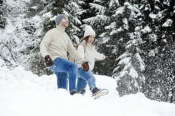 Verspieltes Paar hält sich an den Händen und genießt die Wanderung im Wald bei Schneefall