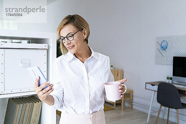Lächelnde Geschäftsfrau hält Kaffeetasse in der Hand  während sie im Büro SMS über ihr Mobiltelefon sendet