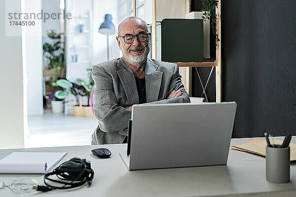 Lächelnder Geschäftsmann sitzt mit verschränkten Armen und schaut im Studio auf den Laptop