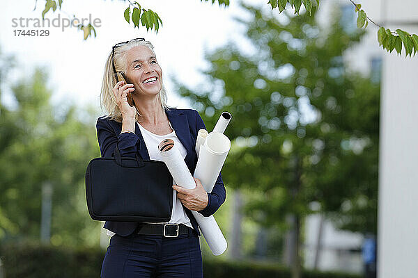 Lächelnde Geschäftsfrau hält Baupläne in der Hand  während sie auf dem Smartphone spricht