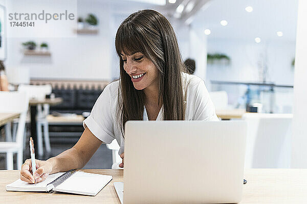 Lächelnde Berufstätige mit Laptop  die am Tisch Tagebuch schreibt