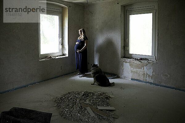 Schwangere Frau mit Hund steht am Fenster ihres renovierten Hauses
