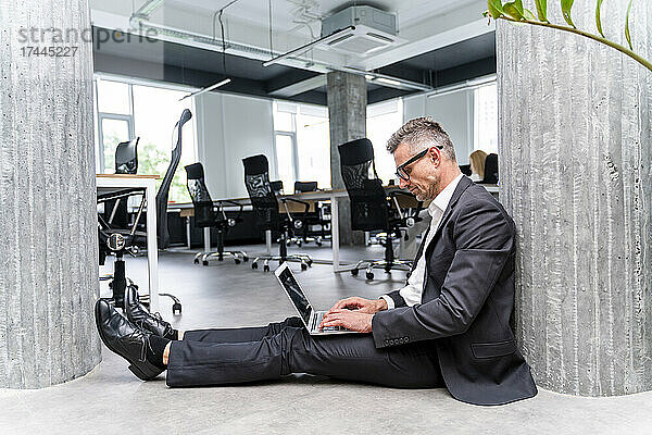 Männlicher Geschäftsprofi benutzt Laptop  während er im Büro auf dem Boden sitzt
