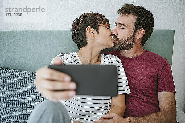 Liebevolles Paar küsst sich  während es zu Hause ein Selfie mit dem Smartphone macht