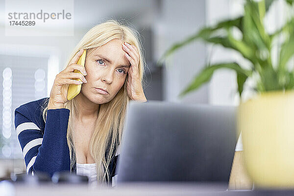 Traurige Geschäftsfrau mit Kopf in der Hand  die im Heimbüro auf dem Smartphone spricht