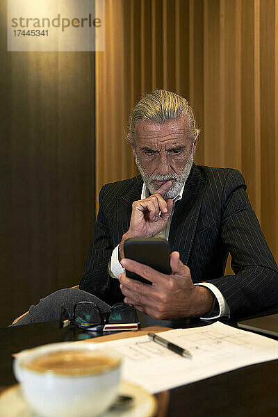Besorgter Geschäftsmann schaut auf sein Smartphone  während er am Schreibtisch im Hotel sitzt