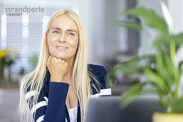 Lächelnde blonde Freiberuflerin mit der Hand am Kinn im Heimbüro