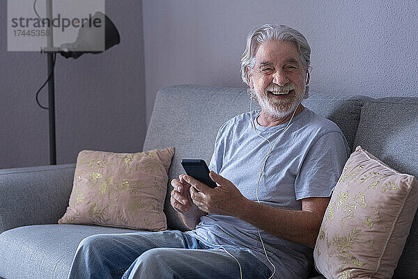 Glücklicher Mann mit Mobiltelefon und In-Ear-Kopfhörern  der zu Hause sitzt