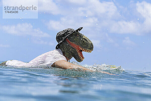 Mann mit Dinosauriermaske schwimmt mit Surfbrett im Meer