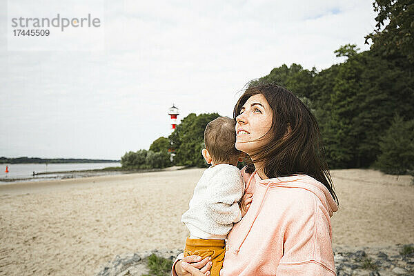Frau schaut nach oben  während sie ihren Sohn am Strand trägt
