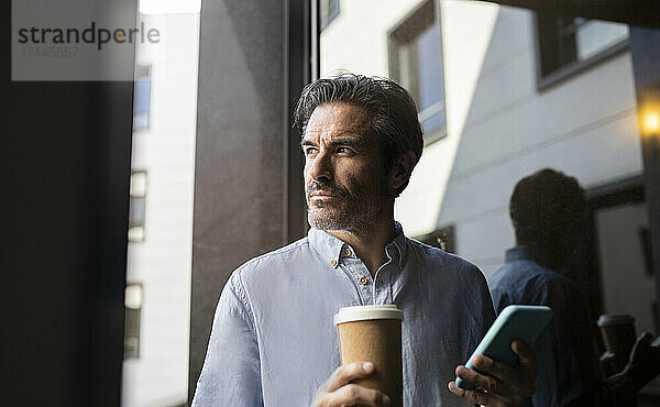 Männlicher Freiberufler hält Mobiltelefon und Einwegkaffeetasse am Fenster
