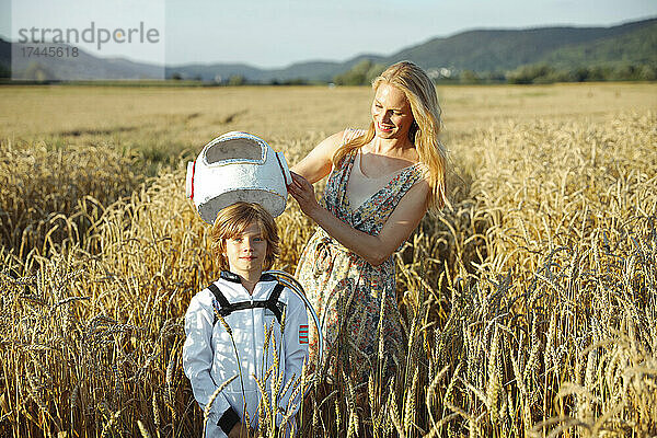 Glückliche Mutter hält Weltraumhelm in der Hand  während sie mit ihrem Sohn auf dem Feld steht