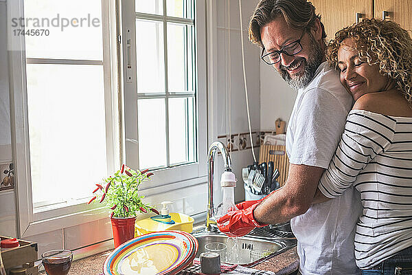 Lächelnde Frau umarmt Mann  der zu Hause in der Küche Geschirr putzt