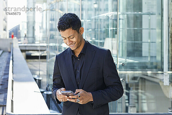 Lächelnder männlicher Berufstätiger  der sein Mobiltelefon benutzt  während er vor dem Bürogebäude in der Stadt steht