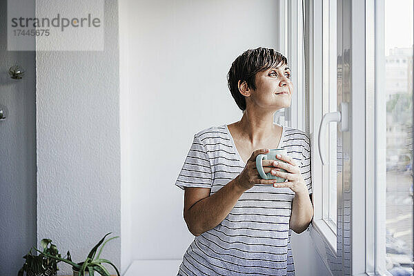 Frau hält Tasse in der Hand  während sie zu Hause durch das Fenster schaut