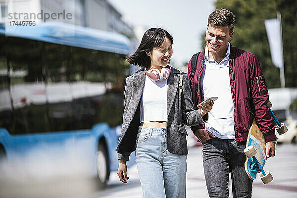 Lächelnde männliche und weibliche Freunde teilen ihr Mobiltelefon  während sie an sonnigen Tagen spazieren gehen