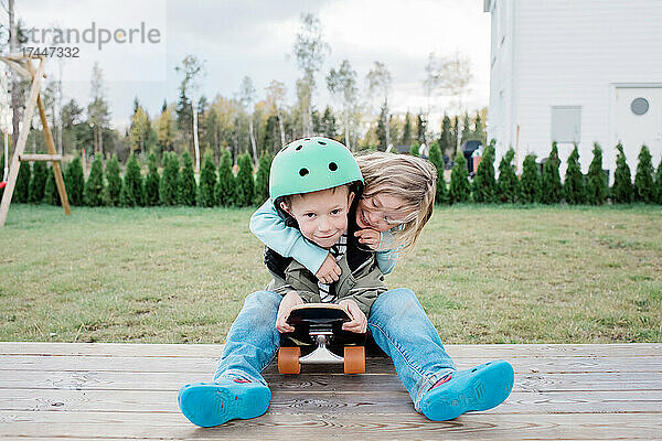 Schwester umarmt ihren Bruder  während sie draußen auf einem Skateboard spielt