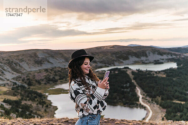 Frau steht mit Handy in einer Landschaft mit einem See und Bergen