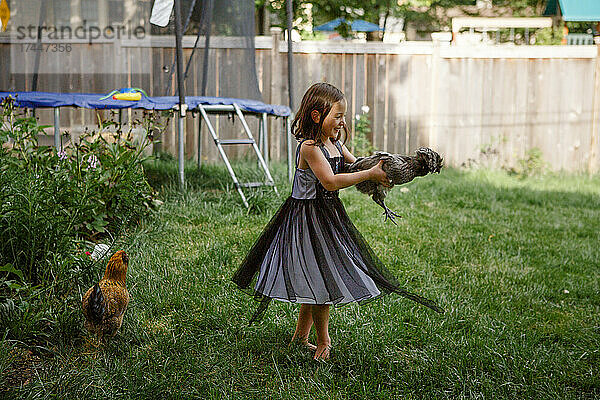 Ein kleines lachendes Mädchen hält ein Huhn und dreht sich im Hinterhofgarten