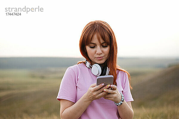 Junge rothaarige Frau mit weißen Kopfhörern schaut in das Smartphone