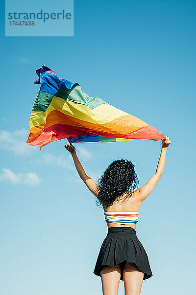 Junge Frau hält eine LGBT-Flagge von hinten. Bekleidet mit einem farbenfrohen Oberteil