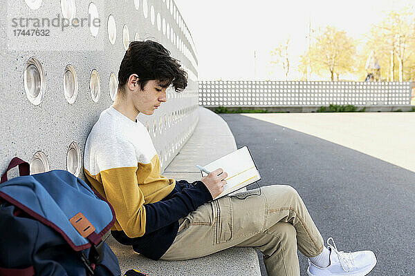 Junger Mann schreibt Notizen in ein Notizbuch und sitzt auf einer Bank