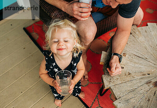 Blondes Kleinkindmädchen trinkt glücklich aus der Tasse  während Papa zusieht