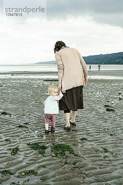 Ein junges Mädchen blickt zurück  während sie am Strand die Hand ihrer Mutter hält