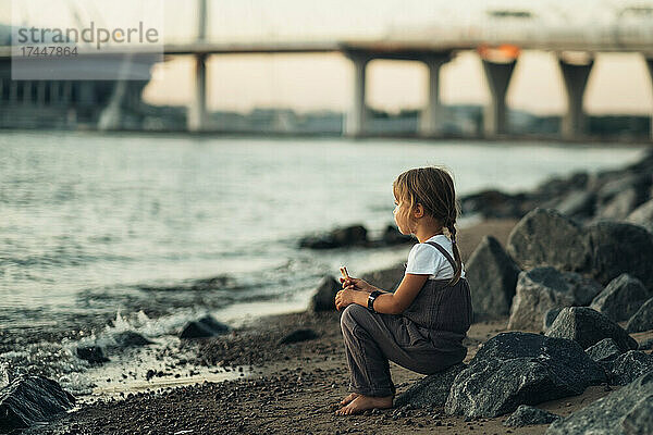 Nettes kleines Mädchen  das bei Sonnenuntergang am Ufer der Bucht sitzt.