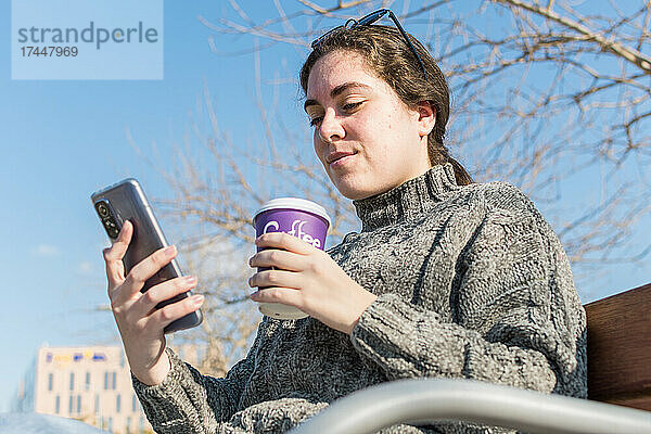 Unteransicht einer jungen Frau  die ein Mobiltelefon benutzt und einen Kaffee in der Hand hält.