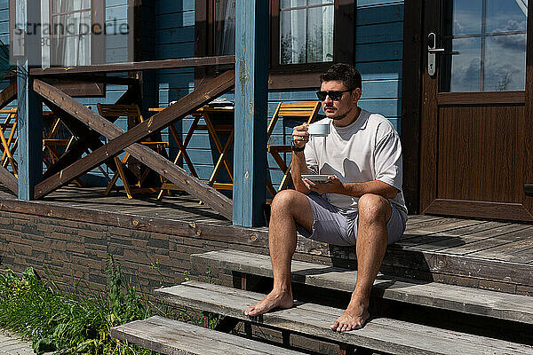 Ein junger Mann sitzt mit einer Tasse Kaffee auf der Veranda eines Hauses