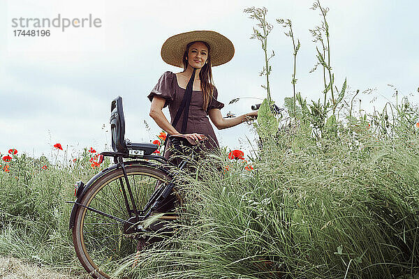 Frau mit Fahrrad steht zwischen Mohnblumenfeld vor dem Himmel