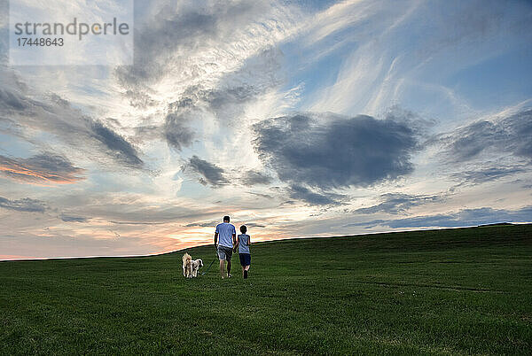 Vater und Sohn gehen bei Sonnenuntergang gemeinsam mit einem Hund einen grasbewachsenen Hügel hinauf.