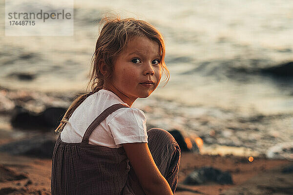 Porträt eines kleinen Mädchens  das bei Sonnenuntergang am Ufer der Bucht sitzt.