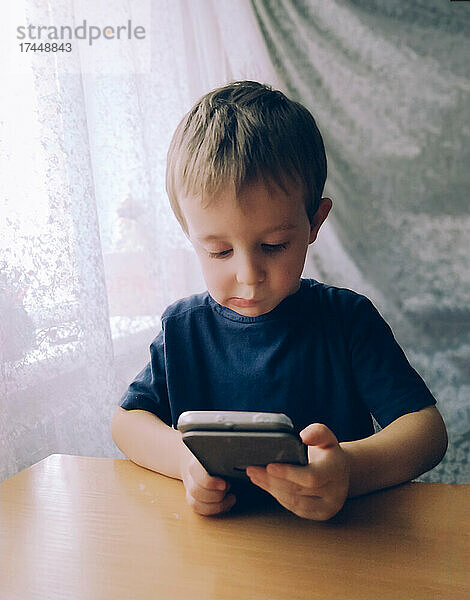 Kleiner Junge schaut drinnen an einem Tisch nachdenklich in ein Smartphone
