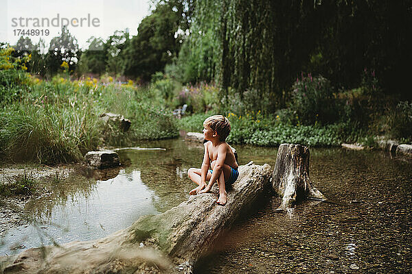 Kleiner Junge sitzt im Sommer auf einem Baumstamm im Wasser und sieht nachdenklich aus