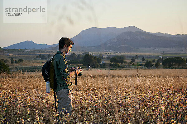 Fotograf mit Rucksack hält bei Sonnenuntergang eine Kamera auf einer Wiese