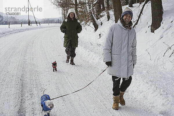 Frauen lachen beim Spazierengehen mit Hunden in Schneejacken auf der gefrorenen Winterlandstraße