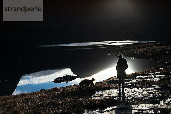 Silhouette einer Frau und eines Hundes vor der Spiegelung der Berge im See