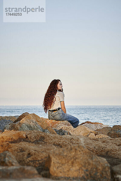 Junge rothaarige Frau blickt auf das Meer  während sie auf Felsen sitzt
