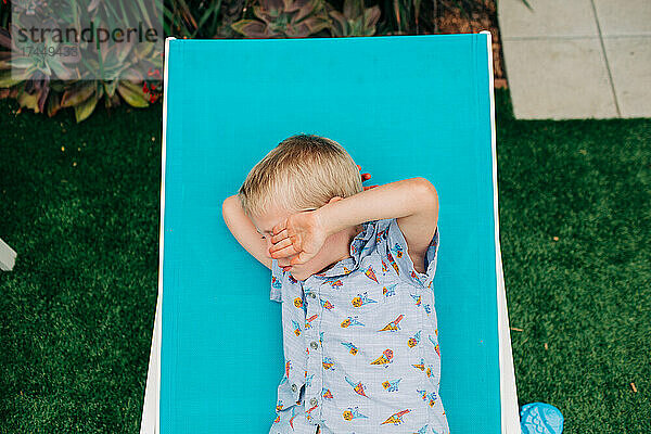 Der blonde Junge schützt seine Augen vor der Sonne  während er im Stuhl faulenzt