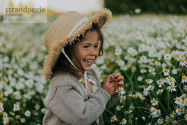 Kleines Mädchen mit Strohhut steht in einem Feld voller Gänseblümchen