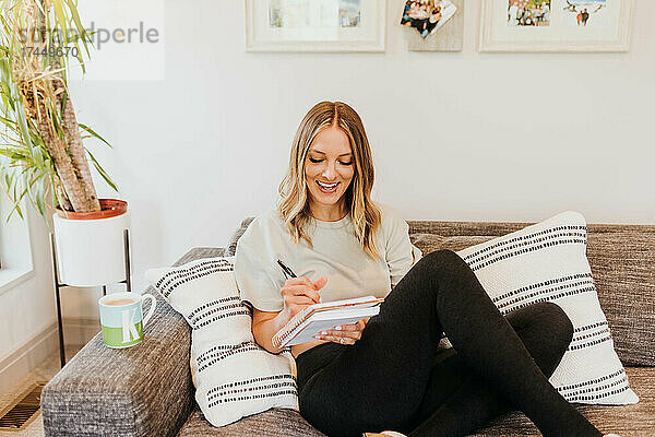 Weitwinkelaufnahme einer lächelnden Frau  die auf der Couch sitzt und in ein Notizbuch schreibt