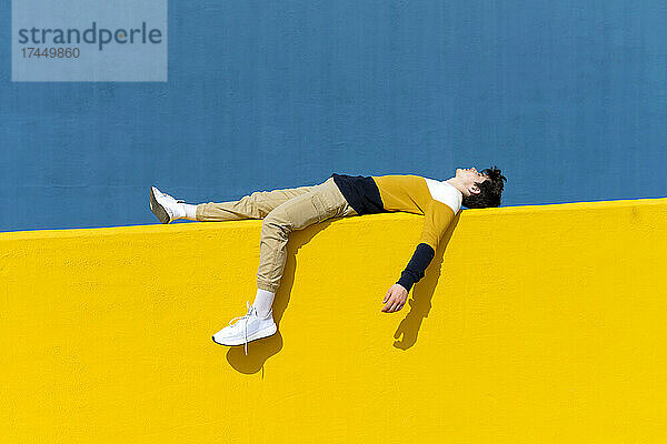 Junger Mann entspannt sich auf gelber Wand vor blauer Wand