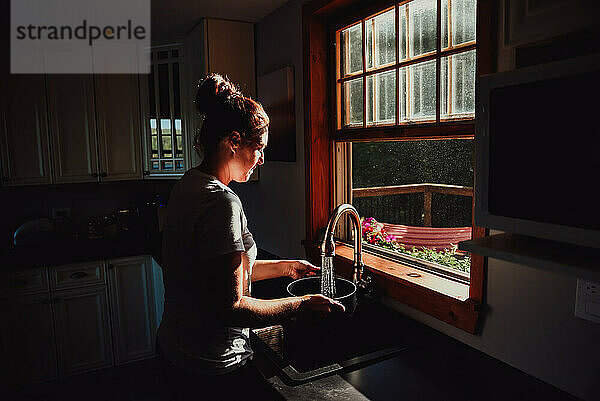 Frau füllt einen Topf am Waschbecken unter einem Fenster in einer dunklen Küche.