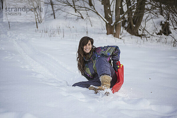 Glückliche Frau im Schneeanzug lächelt  während sie auf der Rodel den Hügel hinunterfährt