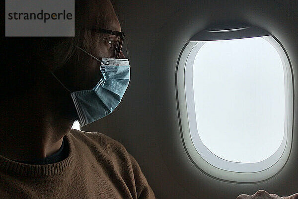 Der Mann trägt eine Gesichtsmaske und blickt durch ein Flugzeugfenster in den Himmel  Pandemie