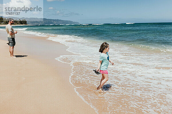 Junges Mädchen geht an einem Strand auf Oahu in Richtung Meer  während Papa zusieht
