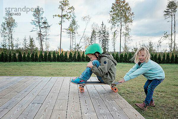 Schwester schiebt ihren Bruder auf einem Skateboard beim Spielen im Garten